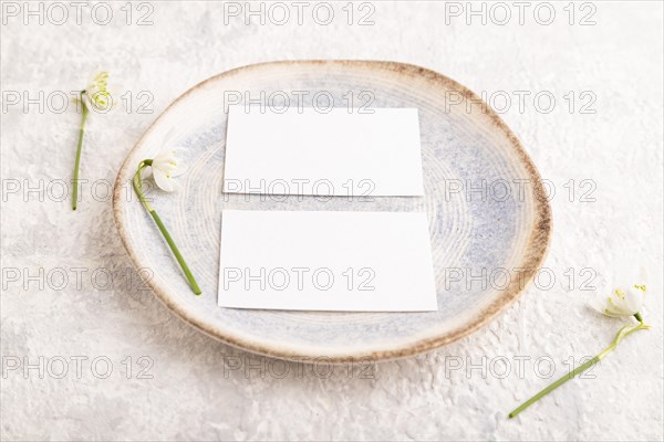 White paper invitation card, mockup with galanthus snowdrop flowers on ceramic plate and gray concrete background. Blank, side view, still life, copy space, wedding invitation