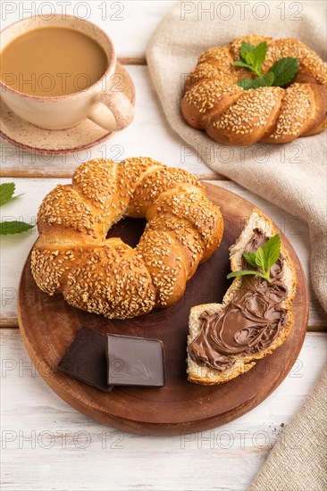 Homemade sweet bun with chocolate cream and cup of coffee on a white wooden background and linen textile. side view, close up