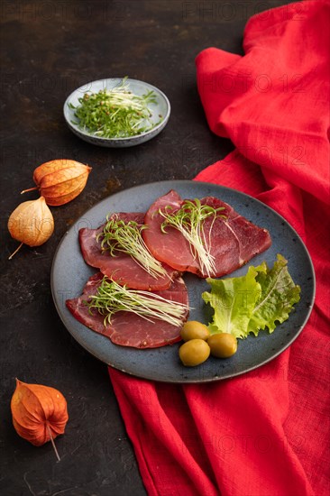 Slices of smoked salted meat with cilantro microgreen on black concrete background and red textile. Side view, close up