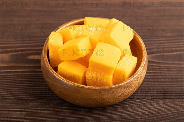 Dried and candied mango cubes in wooden bowls on brown wooden textured background. Side view, close up, vegan, vegetarian food concept