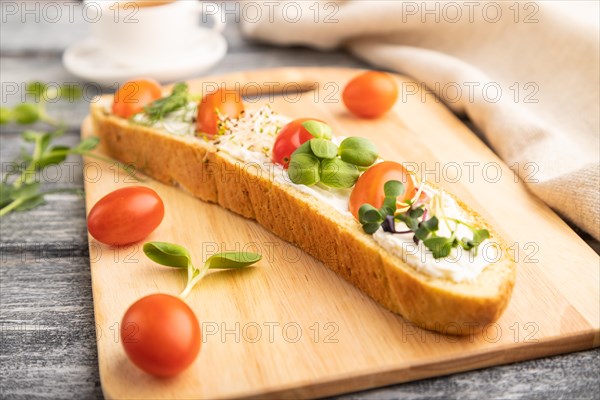 Long white bread sandwich with cream cheese, tomatoes and microgreen on gray wooden background and linen textile. side view, close up, selective focus