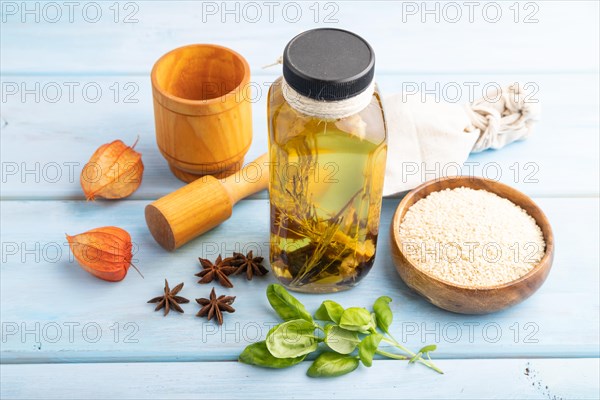 Sunflower oil in a glass jar with various herbs and spices, sesame, rosemary, star anise, basil on a blue wooden background. Side view, close up