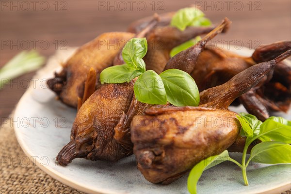 Smoked quails with herbs and spices on a ceramic plate with linen textile on a brown wooden background. Side view, close up