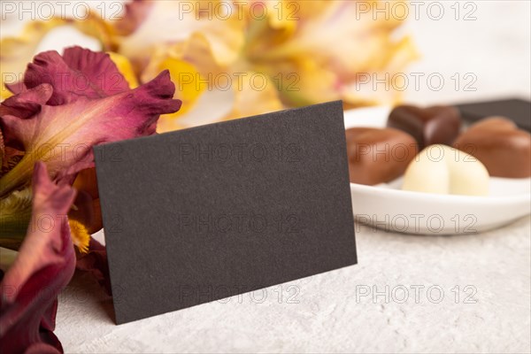 Black business card with chocolate candies and iris flowers on gray concrete background. side view, copy space, still life. Breakfast, morning, spring concept