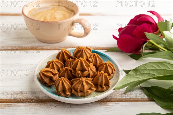 Homemade soft caramel fudge candies on blue plate and cup of coffee on white wooden background, peony flower decoration. side view, close up