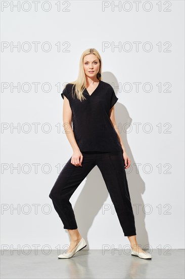 A fashion model wearing a black cotton outfit strikes a dynamic pose against a white wall, showcasing contemporary style and elegance