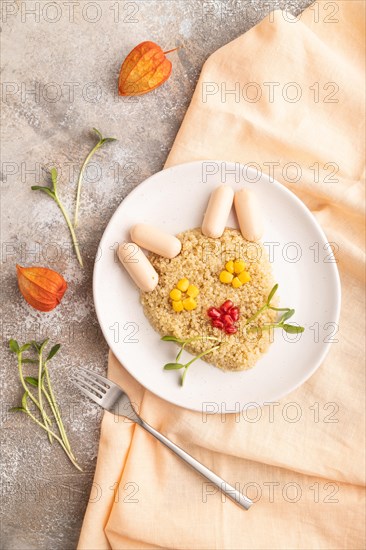 Funny mixed quinoa porridge, sweet corn, pomegranate seeds and small sausages in form of cat face on brown concrete background and orange textile. Top view, flat lay. Food for children, healthy food concept