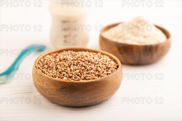 Powdered milk and buckwheat baby food mix, infant formula, pacifier, bottle, spoon on white wooden background. Side view, close up, selective focus, artificial feeding concept