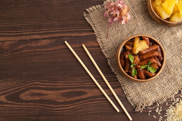 Tteokbokki or Topokki, fried rice cake stick, popular Korean street food with spicy jjajang sauce and pineapple on brown wooden background and linen textile. Top view, flat lay, copy space