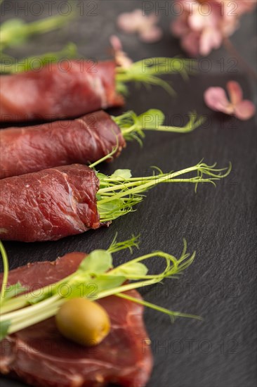 Slices of smoked salted meat with green pea microgreen on black concrete background. Side view, close up, selective focus