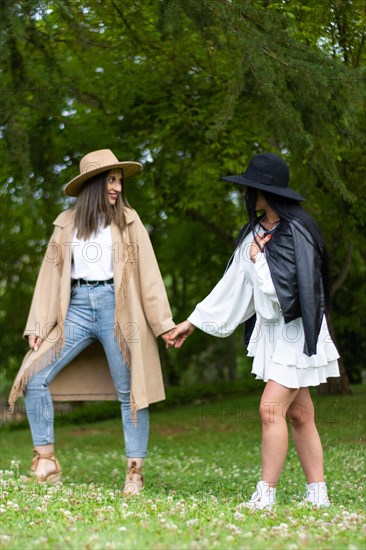 Front view of a stylish lesbian couple in hat holding hands and walking in the park while looking at each other