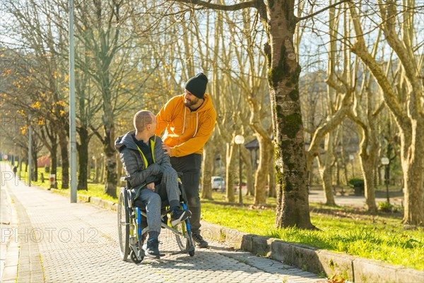 Man pushing the wheelchair of a disabled friend along the path of a park