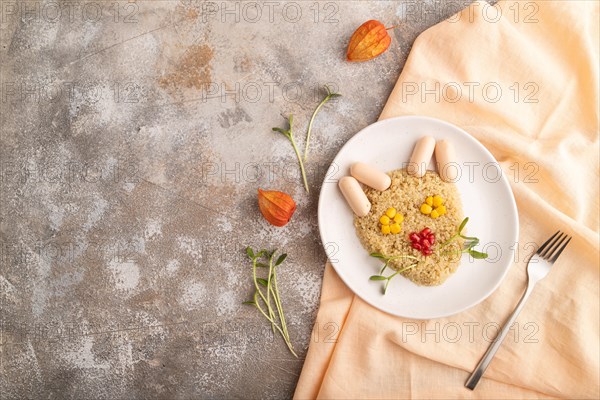 Funny mixed quinoa porridge, sweet corn, pomegranate seeds and small sausages in form of cat face on brown concrete background and orange textile. Top view, flat lay, copy space. Food for children, healthy food concept
