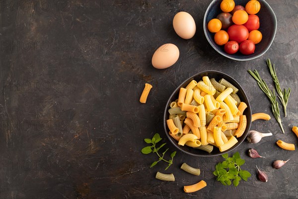 Rigatoni colored raw pasta with tomato, eggs, spices, herbs on black concrete background. Top view, flat lay, copy space