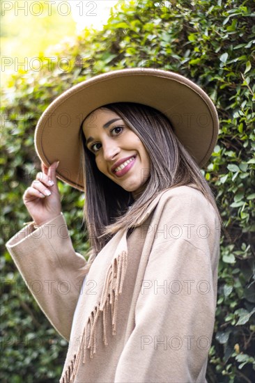 Front vertical view of a young woman in black leather jacket looking at the camera and holding her hat with green leaves in the background outdoors