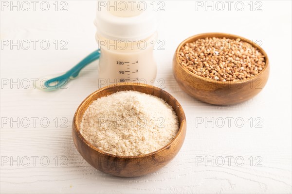 Powdered milk and buckwheat baby food mix, infant formula, pacifier, bottle, spoon on white wooden background. Side view, close up, artificial feeding concept