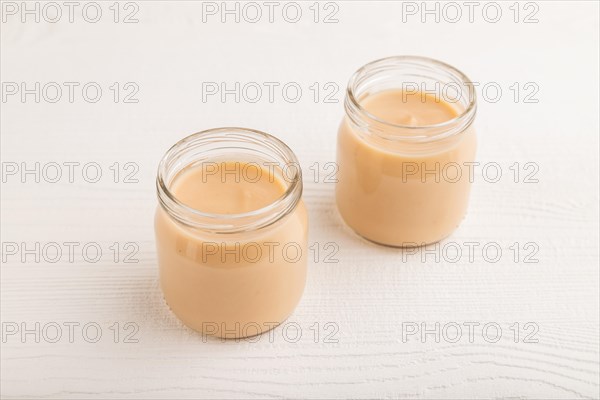 Baby puree with fruits mix, apple, banana infant formula in glass jar on white wooden background. Side view, close up, artificial feeding concept