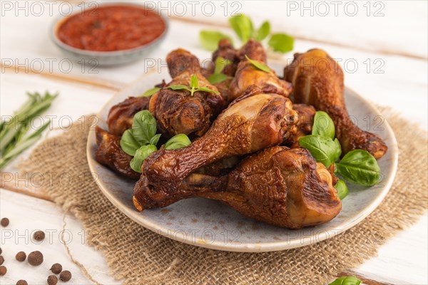 Smoked chicken legs with herbs and spices on a ceramic plate with linen textile on a white wooden background. Side view, close up, selective focus