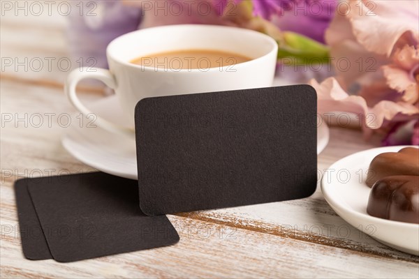 Black business card with cup of cioffee, chocolate candies and iris flowers on white wooden background. side view, copy space, still life. Breakfast, morning, spring concept