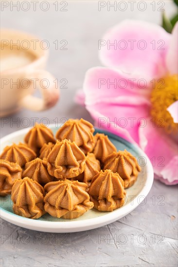 Homemade soft caramel fudge candies on blue plate and cup of coffee on gray concrete background, peony flower decoration. side view, close up, selective focus