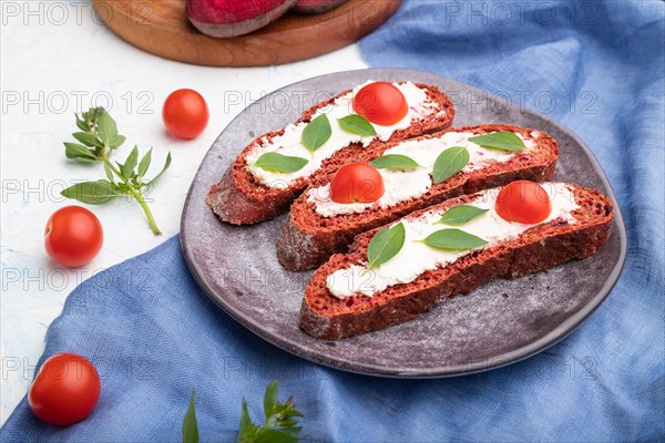 Red beet bread sandwiches with cream cheese and tomatoes on white concrete background and blue linen textile. Side view, close up