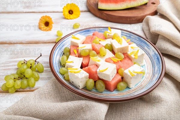 Vegetarian salad with watermelon, feta cheese, and grapes on blue ceramic plate on white wooden background and linen textile. Side view, close up