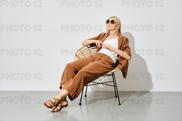 A woman in a loose-fitting suit sits relaxed on a wicker chair in a bright room