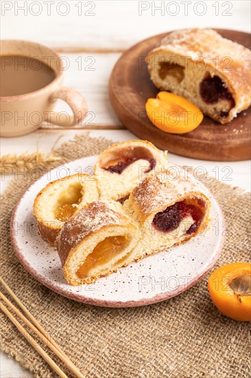 Homemade sweet bun with apricot jam and cup of coffee on white wooden background and linen textile. side view, close up