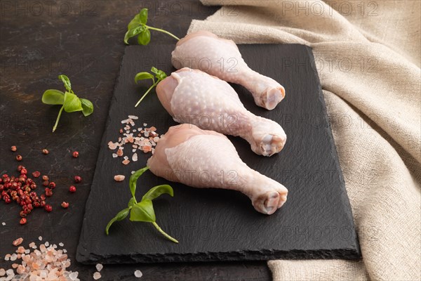 Raw chicken legs with herbs and spices on a black slate cutting board on a black concrete background and linen textile. Side view, close up