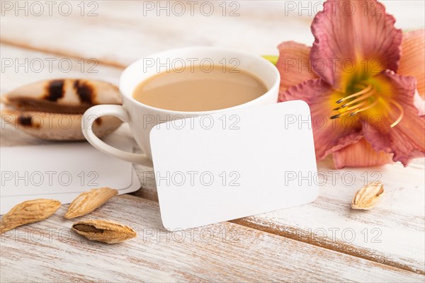 White paper business card mockup with purple day-lily flower and cup of coffee on white wooden background. Blank, side view, copy space, still life. spring concept