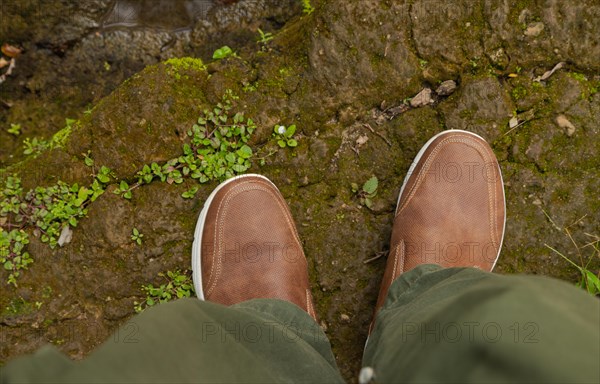 Man feets in hiking leather shoes in outdoors. Top View of shoes on rocky srones near river waterfall, travelling concept