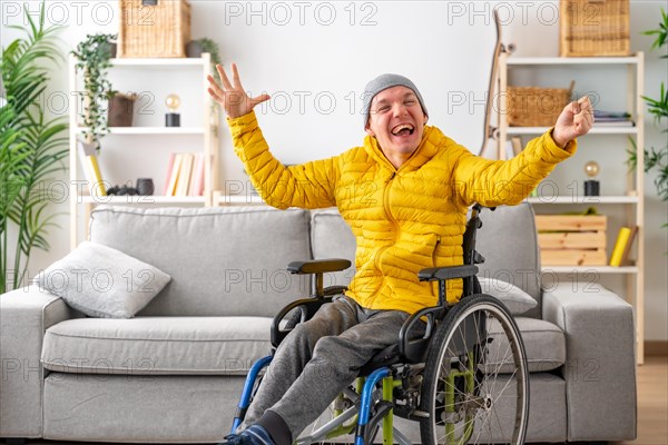 Happy and expressive disabled man in wheelchair raising hands in joy at home