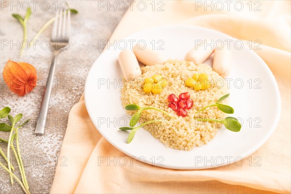 Funny mixed quinoa porridge, sweet corn, pomegranate seeds and small sausages in form of cat face on brown concrete background and orange textile. Side view, selective focus, food for children, healthy food concept