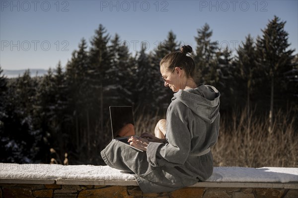 Woman in a bathrobe sits on a snowy stone wall in nature and works on her laptop