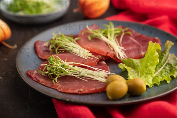 Slices of smoked salted meat with cilantro microgreen on black concrete background and red textile. Side view, close up, selective focus
