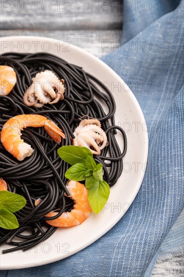 Black cuttlefish ink pasta with shrimps or prawns and small octopuses on gray wooden background and blue textile. Side view, close up, selective focus