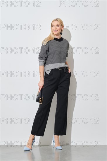 A fashionable woman poses in a striped top and black trousers, accessorized with blue heels and a small purse against a white wall