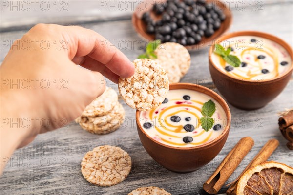 Yoghurt with bilberry and caramel in clay bowl on gray wooden, hand dipping cookies in yogurt, side view, close up, selective focus