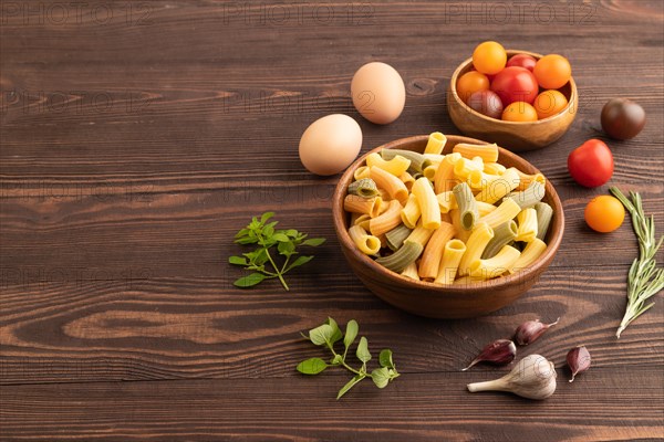 Rigatoni colored raw pasta with tomato, eggs, spices, herbs on brown wooden background. Side view, copy space