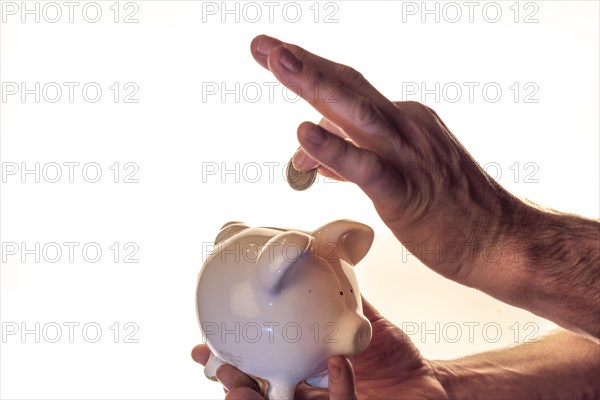 Hand of a man holding a white piggy bank and putting a coin into it, white background, studio shot, Germany, Europe
