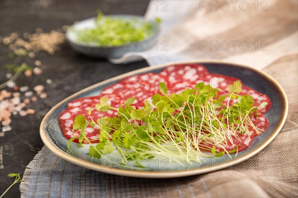 Slices of smoked cervelat salami sausage with spinach microgreen, salt and pepper on black concrete background and beige textile. Side view, close up, selective focus