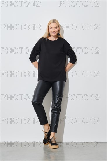 Smiling woman in black pullover and leather trousers leaning against white wall