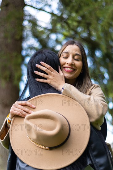 Vertical image of a happy woman hugging her friend with eyes closed in the park while holding her hat