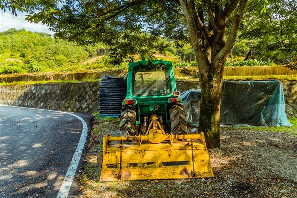 Green tractor with yellow cutting attachment parked under a large shade tree next to a paved road in South Korea