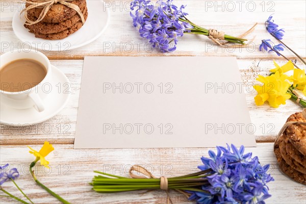 Gray paper sheet with oatmeal cookies, spring snowdrop flowers bluebells, narcissus and cup of coffee on white wooden background. side view, copy space, still life. Breakfast, morning, spring concept