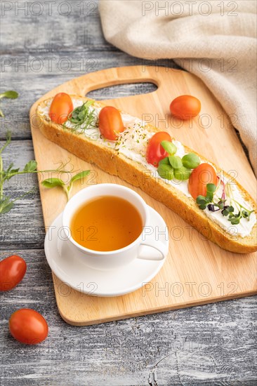 Long white bread sandwich with cream cheese, tomatoes and microgreen on gray wooden background and linen textile. side view, close up