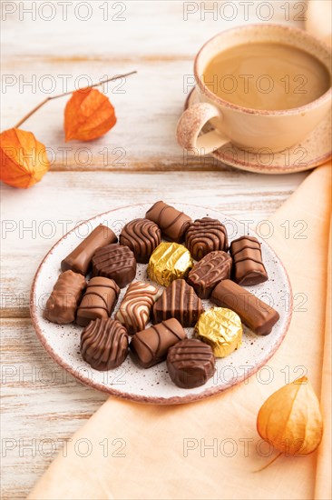 Chocolate candies with cup of coffee and physalis flowers on a white wooden background and orange textile. side view, close up