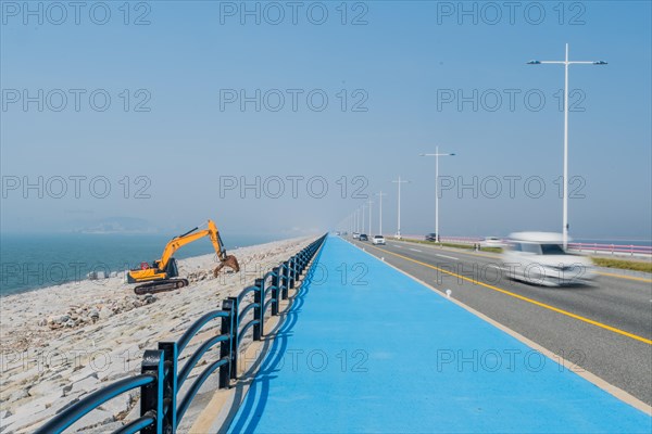 Backhoe sitting on ocean causeway next to blue concrete sidewalk on divided highway with speeding cars passing by. Motion blur caused by slow shutter speed