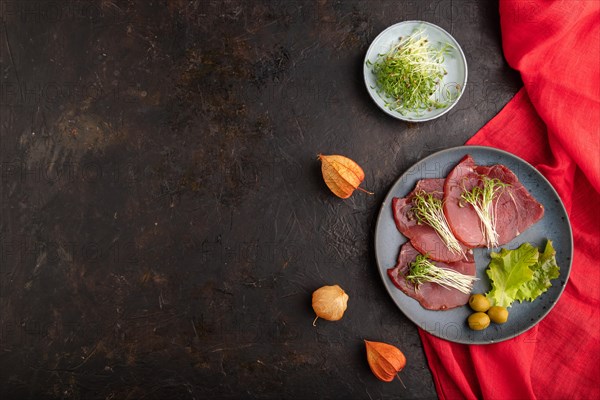 Slices of smoked salted meat with cilantro microgreen on black concrete background and red textile. Top view, flat lay, copy space