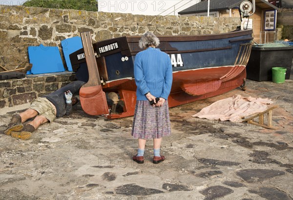 Man painting small fishing boat watched by an elderly woman presumably his mother, Coverack, Cornwall, England, UK